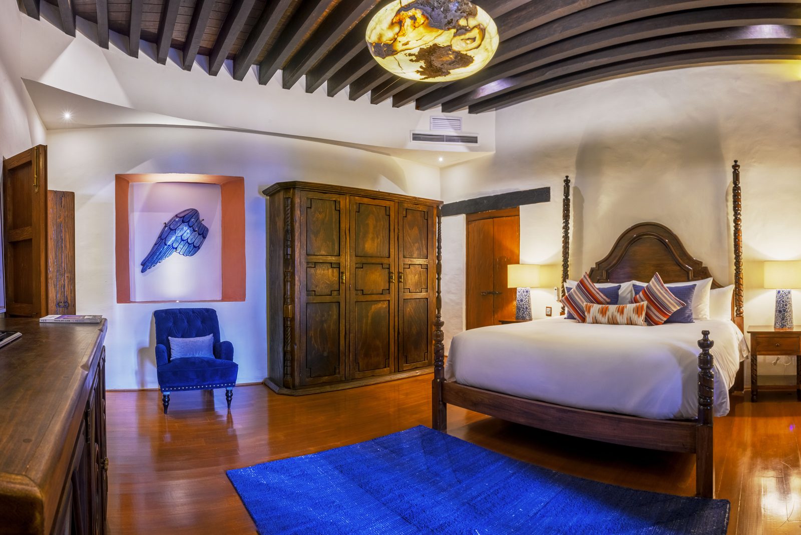 Suite Ángeles con cama de cuatro postes, sillón azul y obra de arte en nicho.